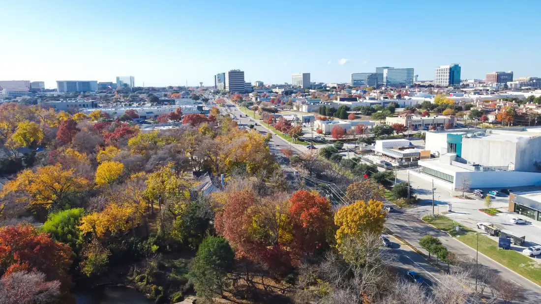 Aerial view of Addison in autumn, with office towers on the skyline above turning tree canopy and the Belt Line commercial strip